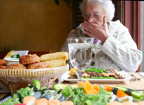 Elderly woman with hand over her mouth and a lot of food in front of her, Heartburn, Acid Reflux, GERD